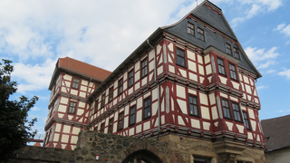 Historisches Fachwerkhaus mit rot-weißer Fassade, vielen Fenstern und dunklem Schieferdach, von unten vor bewölktem Himmel.