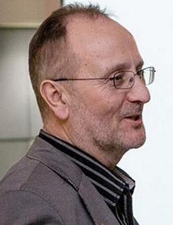 Profil eines glatzköpfigen Mannes mit Brille, kurzem Bart, grauem Sakko und gestreiftem Hemd, lächelnd oder sprechend.