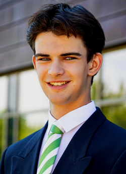 Ein lächelnder junger Mann in einem dunklen Blazer und einer grün gestreiften Krawatte steht vor einem modernen Gebäude.