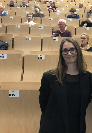 Junge Frau mit Brille lächelt in die Kamera, im Hintergrund ein vollbesetzter Hörsaal mit Reihen von Zuhörern.