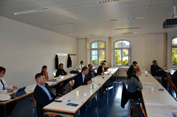 Teilnehmer, teils uniformiert, bei einer Konferenz oder einem Seminar in einem hellen Raum mit großen Fenstern.