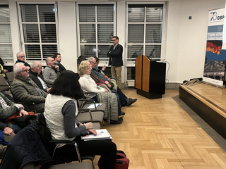 Mann spricht an einem Rednerpult vor Publikum in einem Raum mit einem Banner der 'Gesellschaft für Sicherheitspolitik e.V.'