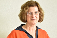 Lächelnde Frau mit lockigen braunen Haaren und Brille, trägt ein dunkelblaues Hemd und einen orangefarbenen Cardigan.