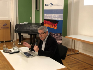 Sprecher gestikuliert mit Mikrofon an Laptop bei Sicherheitspolitik-Veranstaltung; GSP-Banner im Hintergrund.