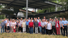 Gruppe Senioren und weitere Personen posieren vor einem grünen, historischen Militärflugzeug, dem 'Fassberg Flyer', unter einem Unterstand.