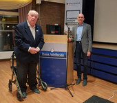 Zwei Männer, einer mit Rollator, der andere kahl, bei einer Veranstaltung neben einem Pult und Reservistenbanner.