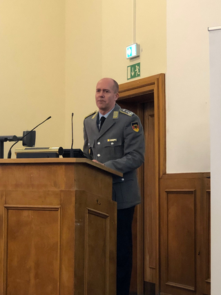 Ein kahler Mann in deutscher Militäruniform steht hinter einem Holzpult mit Mikrofonen und hält eine Präsentation.