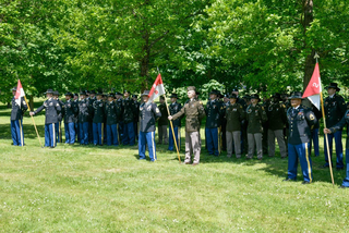 Eine Gruppe Soldaten in verschiedenen Uniformen steht in Formation auf einem sonnigen grünen Rasen mit Bäumen im Hintergrund, einige halten Einheitsfahnen.