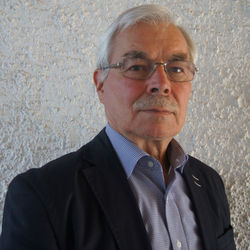 Älterer Mann mit grauem Haar, Brille und Schnurrbart, trägt ein kariertes Hemd und eine dunkle Jacke.