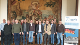 Gruppe von 15 Personen (meist Männer, eine Frau) vor antikem Wandteppich und GSP-Banner.