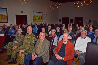 Ein Publikum aus Erwachsenen, darunter ein Mann in Militäruniform, sitzt in Reihen in einem Auditorium und schaut aufmerksam zu.