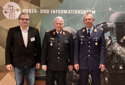 Drei Männer, zwei Militäroffiziere und ein Zivilist, vor einem Banner "CYBER- UND INFORMATIONSRAUM" bei einem Event.