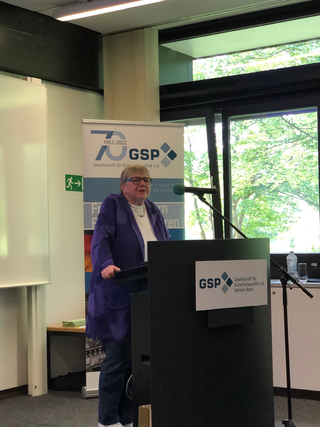 Eine Frau in violetter Jacke spricht an einem Rednerpult mit GSP-Logo, dahinter ein 70-Jahre-Banner und ein Fenster mit Bäumen.