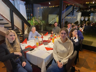 Eine Gruppe von Menschen versammelt an einem Restauranttisch mit Getränken, Kerzen und roten Servietten. Zwei Frauen lächeln in die Kamera.