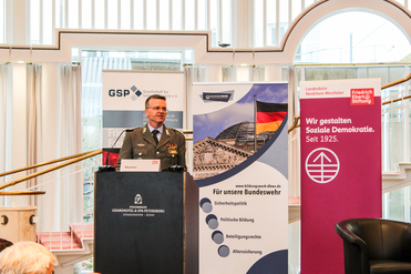 Militäroffizier Wüstner spricht an einem Rednerpult auf einer Konferenz, mit Bannern von Bundeswehr und Stiftungen.