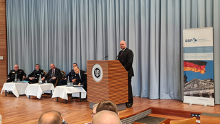 Redner am Pult der Führungsakademie Bundeswehr, im Hintergrund sitzende Offiziere und GSP-Banner mit Deutscher Flagge und Reichstag.