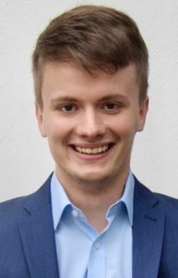 Ein junger Mann im blauen Anzug und hellblauem Hemd lächelt in die Kamera.