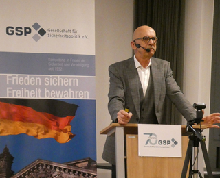 Mann mit Brille und Headset-Mikrofon spricht am Rednerpult vor GSP-Banner mit deutscher Flagge, 70-jähriges Jubiläum.
