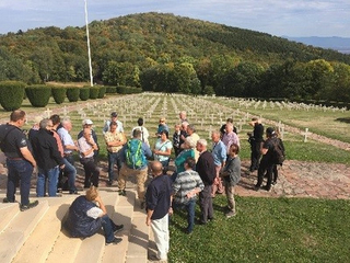 Besuchergruppe auf einem Soldatenfriedhof mit zahlreichen weißen Kreuzen und einem bewaldeten Hügel im Hintergrund.