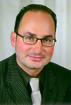 Porträt eines lächelnden Mannes mit kurzen dunklen Haaren, Brille, dunklem Nadelstreifenanzug, dunklem Hemd und heller Krawatte.