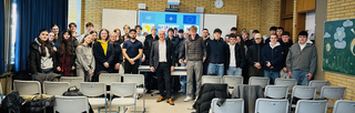 Schüler und Erwachsene in einem Klassenzimmer, mit Logos internationaler Organisationen (UN, NATO, EU, G7) auf Bildschirm.