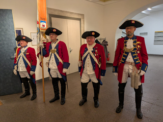 Vier Männer in aufwendigen rot-blauen historischen Militäruniformen mit Dreispitzhüten und hohen Stiefeln, einer hält ein Banner.
