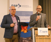 Zwei Männer mit Mikrofonen präsentieren bei einer Veranstaltung der Gesellschaft für Sicherheitspolitik (GSP), GSP-Banner und deutsche Flagge sichtbar.