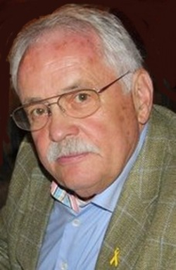 Nahaufnahme eines älteren Mannes mit Brille, Schnurrbart, kariertem Sakko und bunter Fliege, der eine gelbe Schleife trägt.