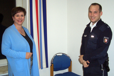 Eine lächelnde Frau und ein lächelnder Polizist in Uniform stehen nebeneinander vor einer Wand mit gestreiftem Vorhang.