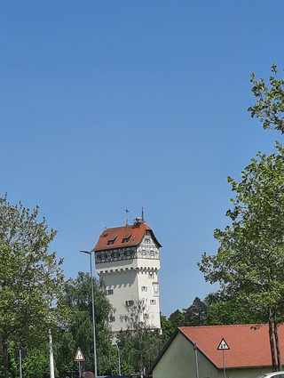 Ein hoher, weißer Wasserturm mit rotem Fachwerk-Dach ragt vor klarem blauem Himmel empor, umrahmt von grünen Bäumen.