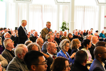 Vielfältiges Publikum lauscht einem Redner auf einer Konferenz. Ein Mann spricht ins Mikrofon, ein anderer filmt.