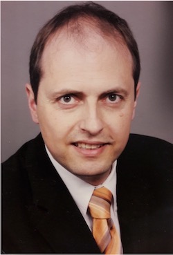 Porträt eines lächelnden Mannes mit kurzen braunen Haaren, der einen dunklen Anzug und eine gestreifte orangefarbene Krawatte trägt.