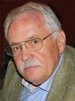 Kopfportrait eines älteren Mannes mit grauem Haar, Brille und Schnurrbart, der einen gemusterten Blazer trägt.