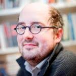 Ein lächelnder Mann mit Brille, Bart und Geheimratsecken blickt in die Kamera. Bücherregale verschwommen im Hintergrund.