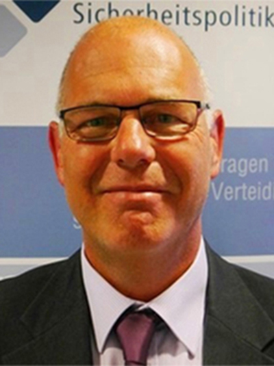 Lächelnder, glatzköpfiger Mann mit Brille, dunklem Anzug und lila Krawatte vor einem Banner über 'Sicherheitspolitik'.