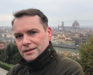 Ein Mann in einer Jacke blickt in die Kamera, im Hintergrund eine Panoramaansicht von Florenz mit dem Dom.